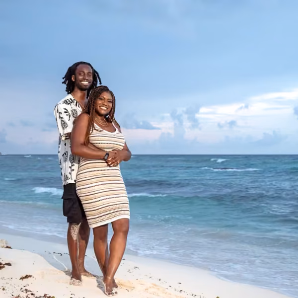 A barefoot couple embraces on a sandy beach at sunset, with blue sky and waves behind them; she wears stripes, he a patterned shirt.