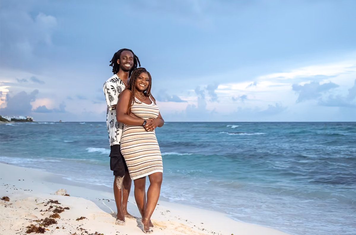A barefoot couple embraces on a sandy beach at sunset, with blue sky and waves behind them; she wears stripes, he a patterned shirt.