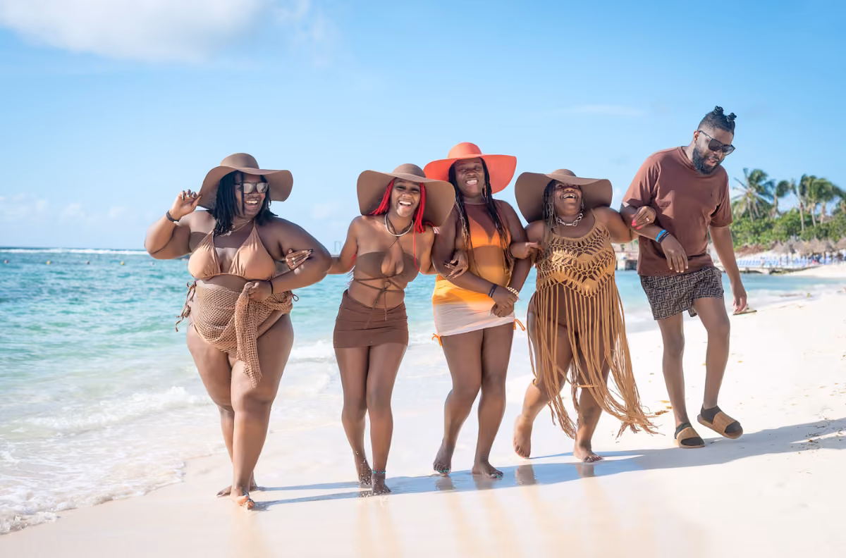 Four women in swimsuits and hats and a man in shorts laugh on a sunny beach as a photographer snaps photos by the blue water.
