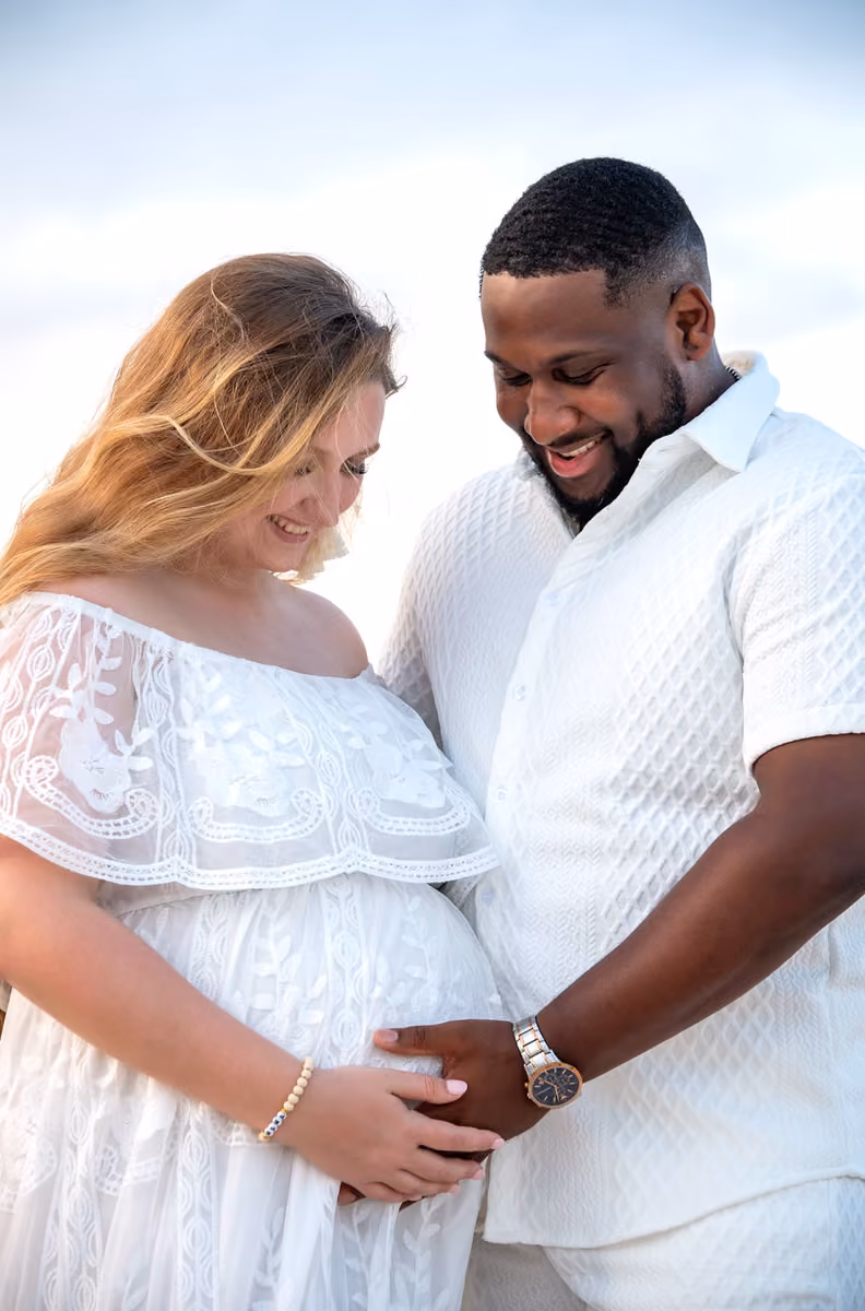A smiling pregnant woman and her partner stand outside in white outfits, gazing at each other as he gently touches her belly.