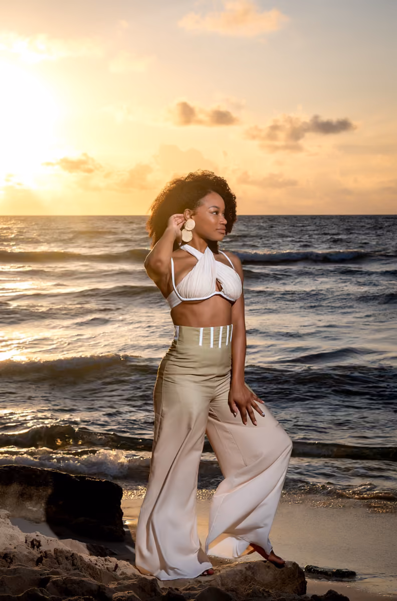 A woman in a white halter top and beige pants poses on the beach at sunset, waves and golden sky behind her.