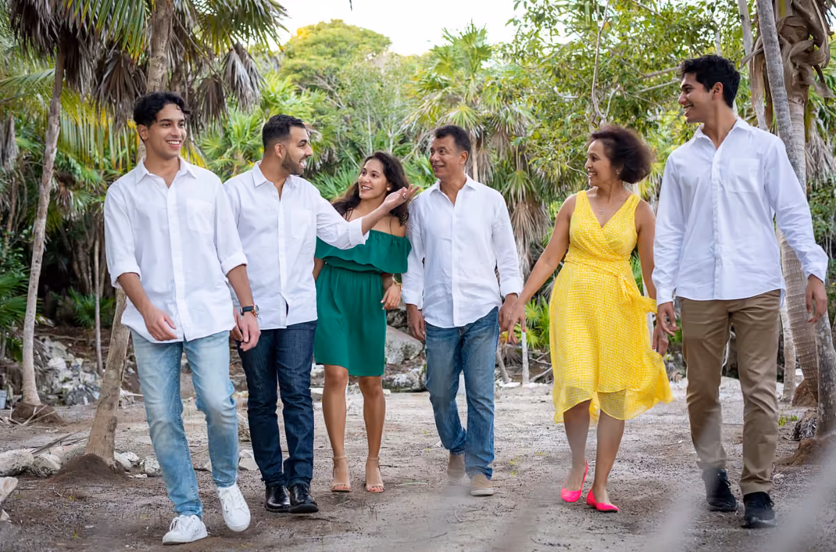 A smiling family of six walks together on a dirt path through lush green trees, enjoying a sunny day outdoors.