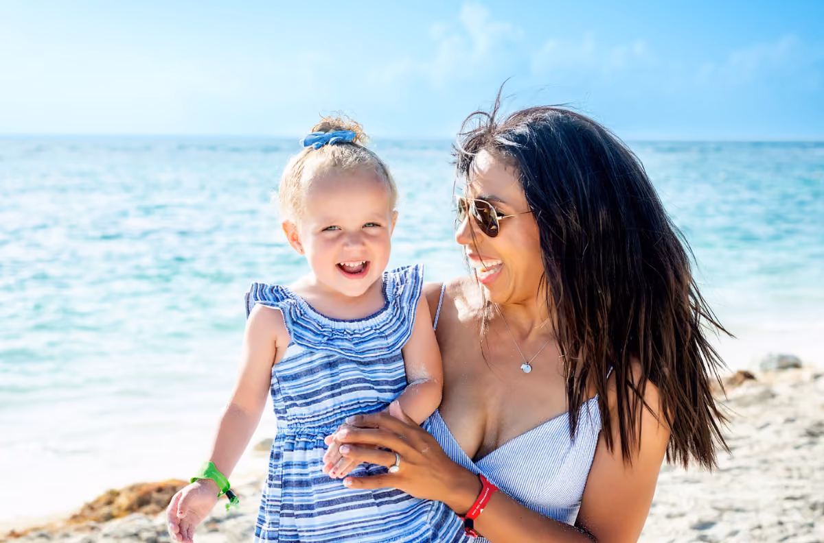 A woman in sunglasses holds a smiling girl on a sunny beach, while someone photographs them with blue ocean and clear sky behind.