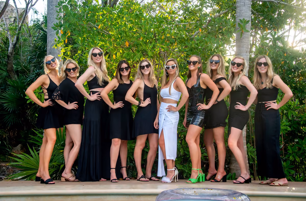 Eleven women pose outdoors, all in sunglasses and black except one in a white dress with green heels, lush foliage behind them.