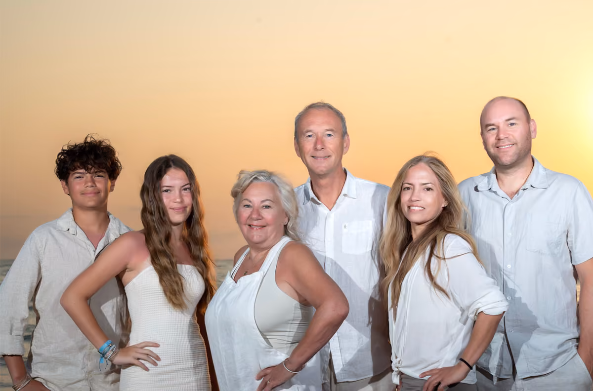 Six people in light clothes smile at sunset, posing outdoors as a photographer captures them against a yellow-orange sky.