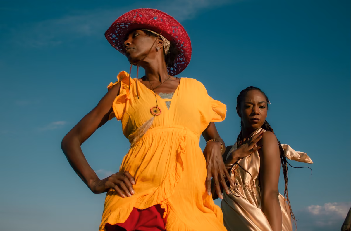 Two women stand outdoors under a blue sky, one in a yellow dress and red hat, the other in beige, both looking different ways.