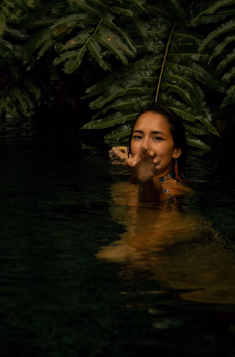 A woman reaches forward, half-submerged in water beneath lush leaves, soft light creating a tranquil, tropical feel.