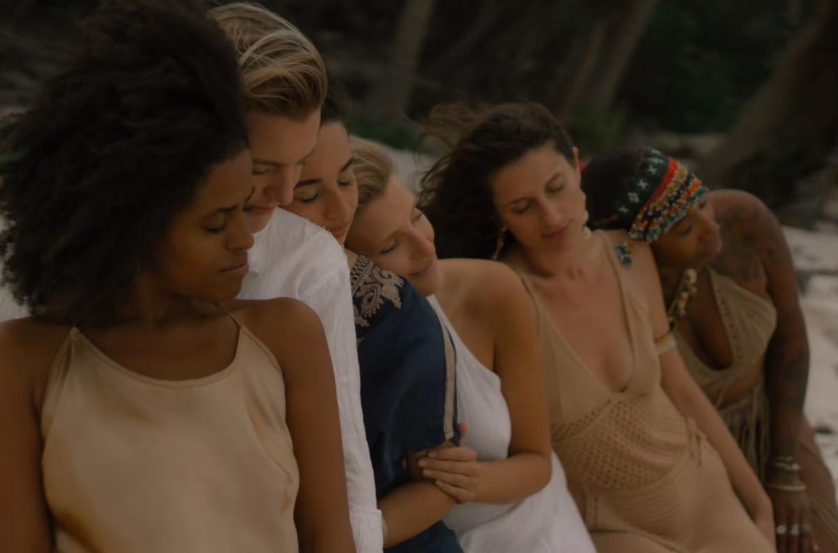 Five women sit closely outdoors, eyes closed and leaning on each other, relaxed in light summer clothes, enjoying a calm moment.