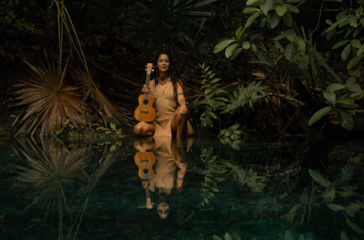 A woman in a tan dress kneels by a clear pond in a lush forest, holding a guitar as her reflection shimmers in the water.