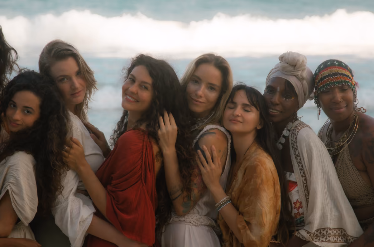 Seven women smile and embrace on a beach, wearing boho outfits, with ocean waves and a clear sky in the background.