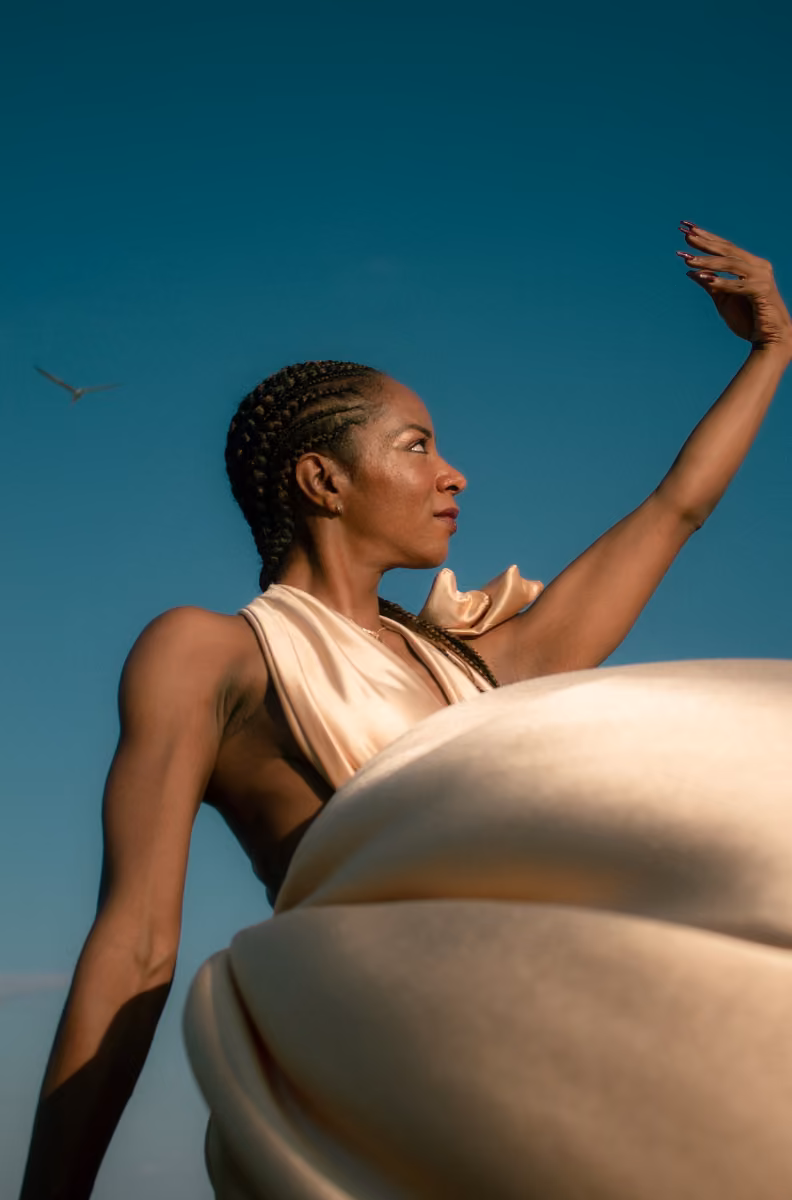 A woman in a flowing beige gown poses gracefully under a clear blue sky, one arm raised, with a distant bird to her left.