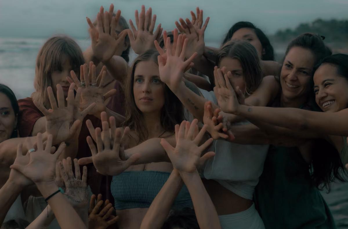 Women stand together outdoors, raising open hands with some dirt on their palms. Gentle sunlight and a beach set a calm mood.