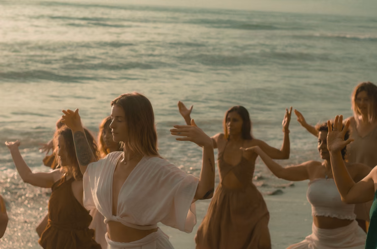 People with eyes closed and arms raised dance or move mindfully on a beach at sunset, waves rolling in behind them.