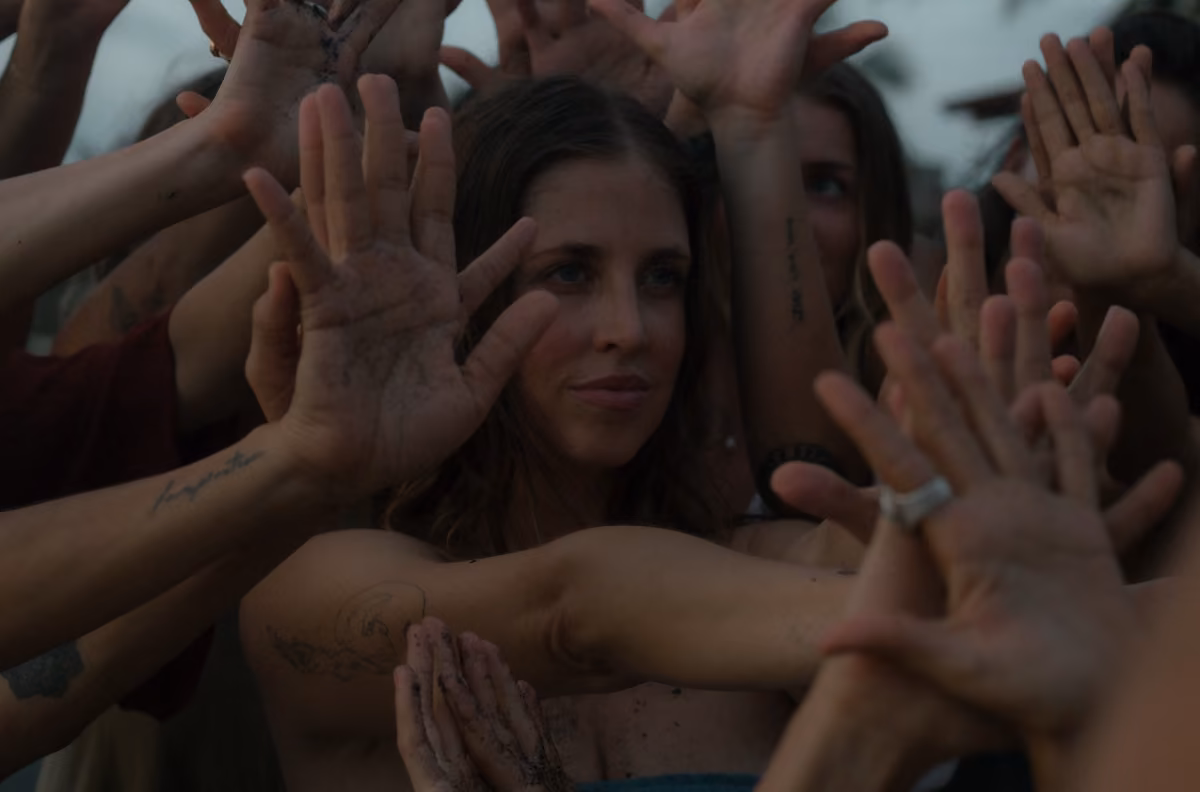 A woman sits calmly as several hands, some dirty, reach toward her in soft light, creating an intimate and communal scene.