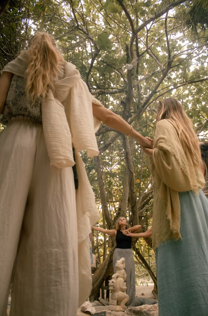 Women stand in a circle outdoors, holding hands under leafy trees as sunlight streams through during a peaceful gathering.