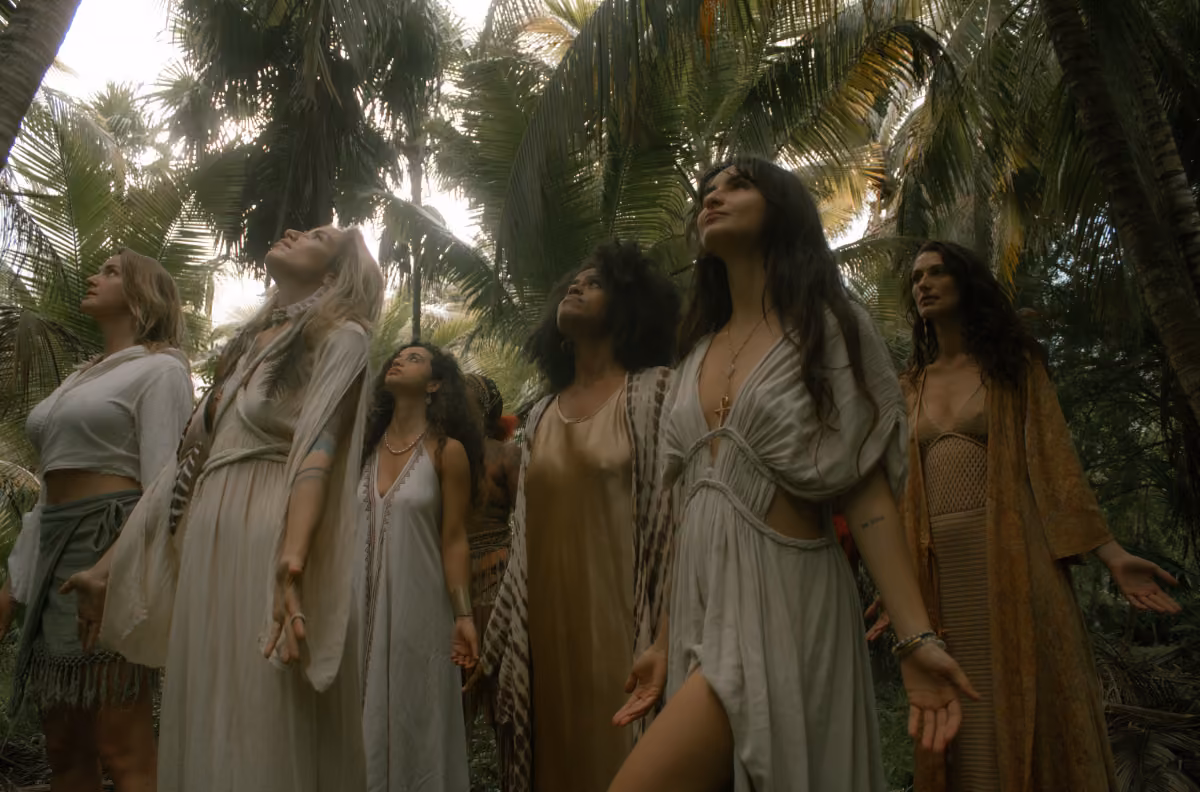 Six women in flowing dresses stand among palm trees in a tropical forest, gazing upward serenely as sunlight filters down.