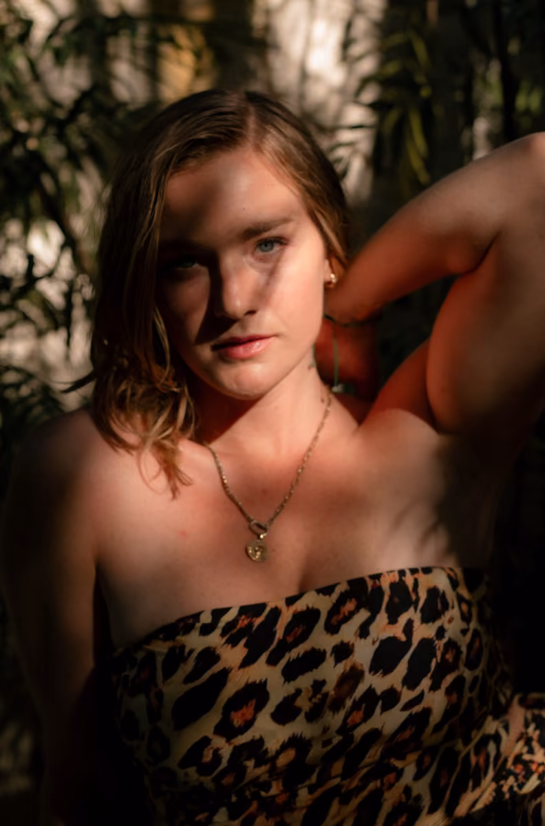A woman in a strapless leopard print top and gold necklace poses with one arm up, sunlight and greenery behind her.