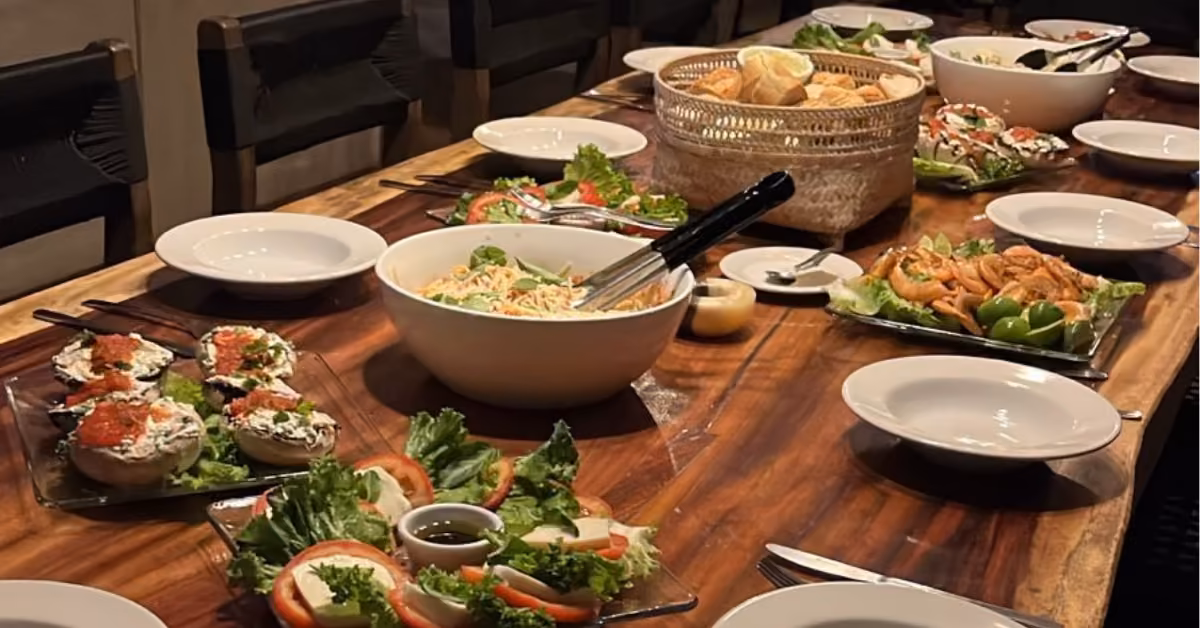 Wooden table set for a meal, with plates, cutlery, salad, bread, pasta, sandwiches, and appetizers arranged for guests.