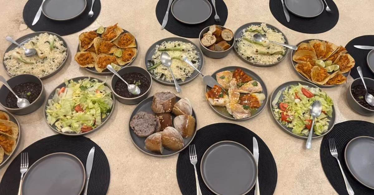 Table set for six with salads, bread rolls, beans, rice, and mains. Utensils rest on black placemats around the dishes.