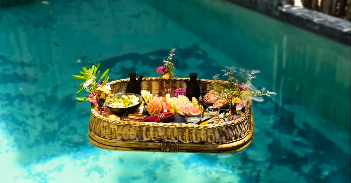 A wicker tray with assorted food and drinks floats on clear pool water, decorated with fresh flowers and greenery.