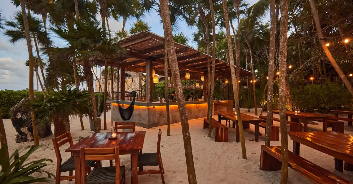 Beachside bar with wooden tables on sand, palm trees, and string lights, creating a cozy dusk atmosphere for gatherings.