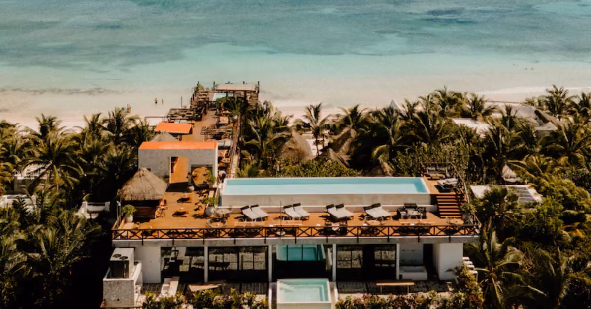 Aerial view of a beachfront resort with palm trees, rooftop pool, sun loungers, and seating overlooking turquoise ocean water.