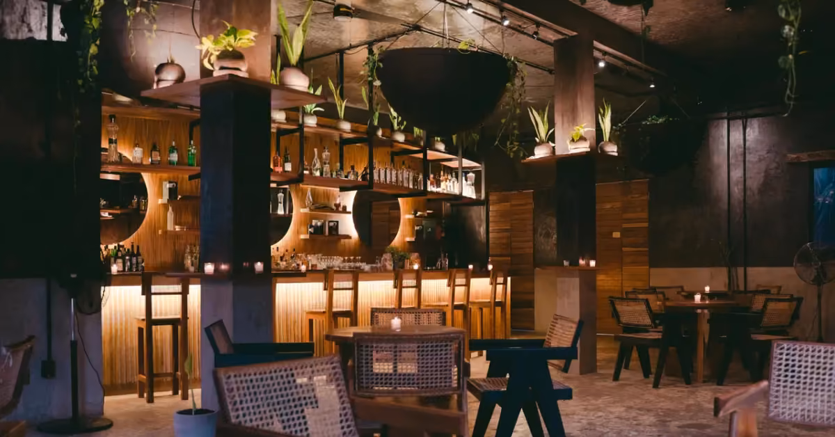 Modern bar with wood furnishings, woven chairs, plants, warm lighting, bottles on shelves, and candles on tables.