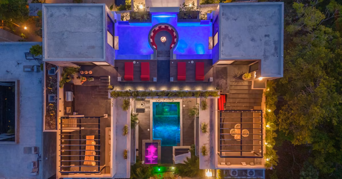 Aerial view of a luxury villa at night with two glowing pools, rooftop lounges, red and white seating, and lush greenery.
