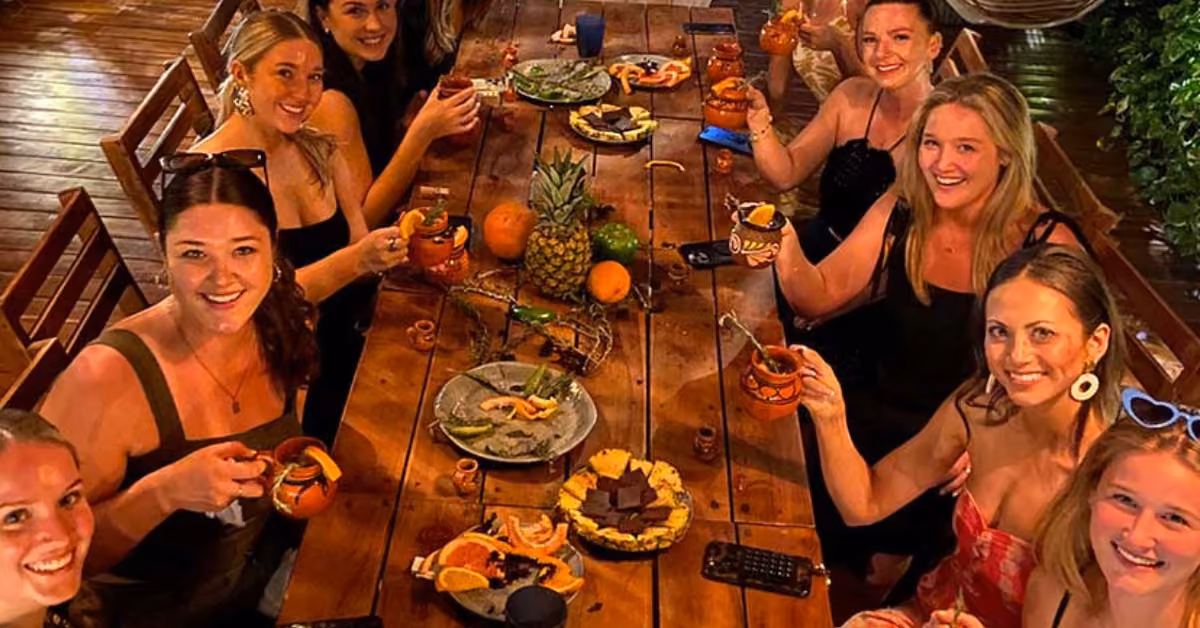 Eight women smile around a wooden table with drinks, fruit, cheese, and festive mugs in a warm, cheerful setting.