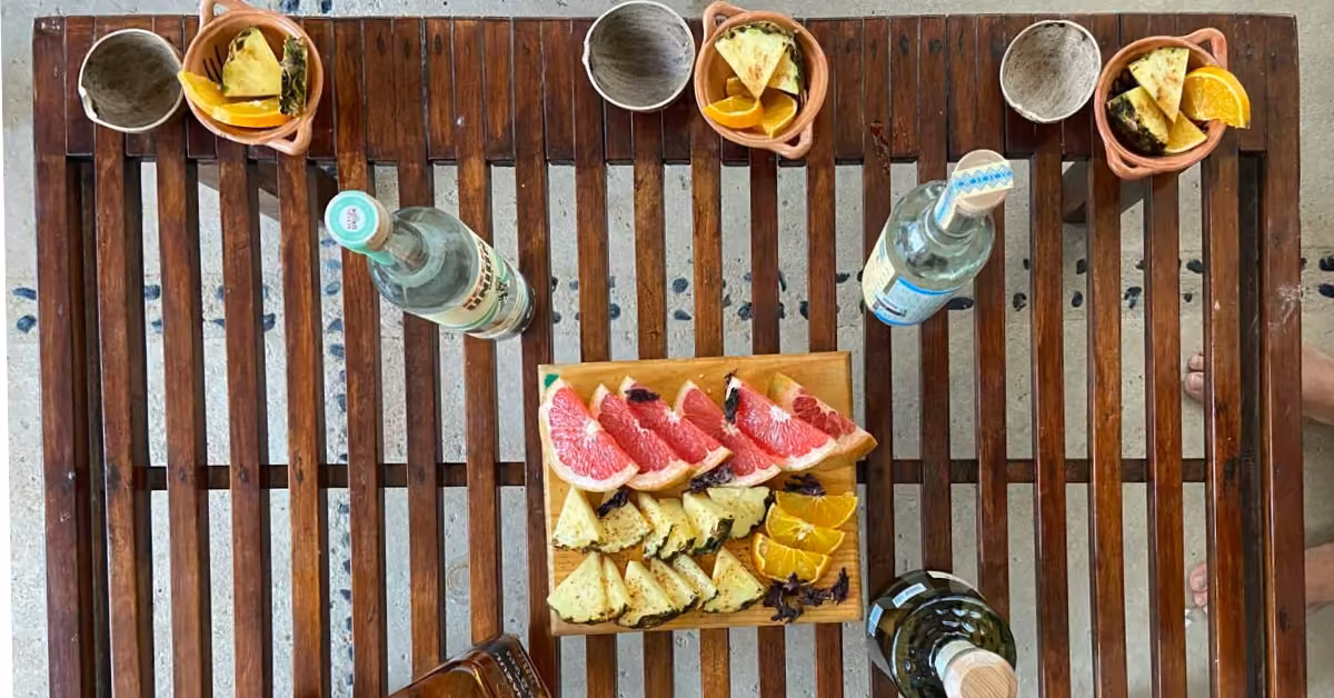 Overhead view of a wooden table with sparkling water bottles, wine, clay cups, and plates of pineapple, orange, and grapefruit.