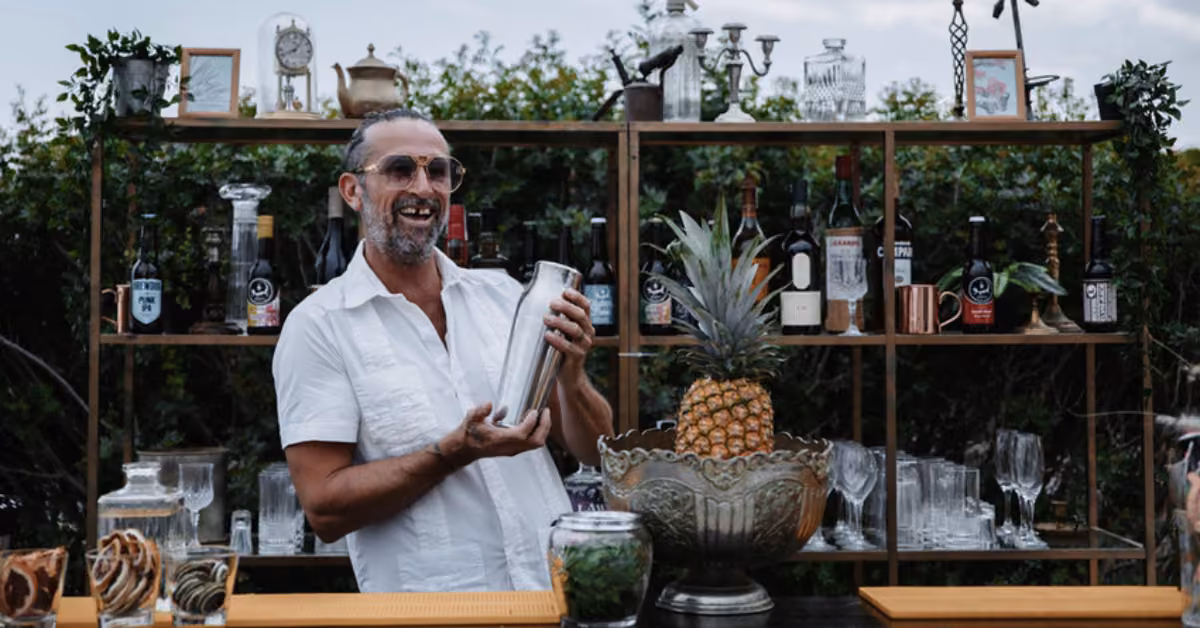 A smiling man in a white shirt shakes a cocktail behind an outdoor bar with bottles, glasses, and pineapple, framed by greenery.