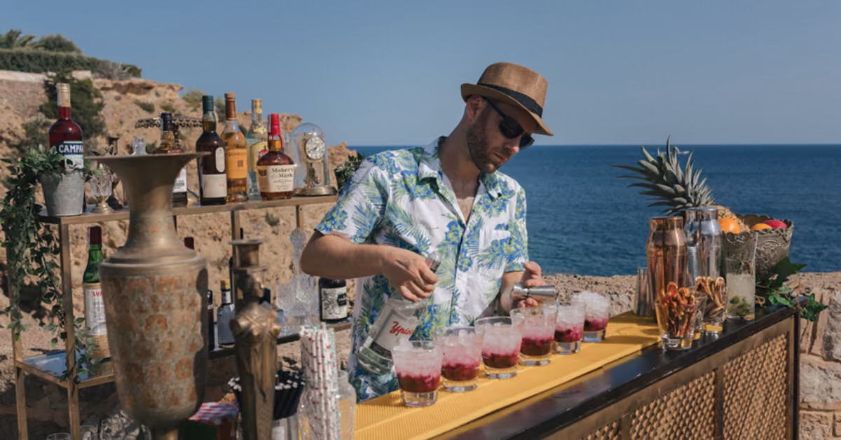 A man in a straw hat and floral shirt makes cocktails at a seaside outdoor bar, fresh fruit and bottles arranged nearby.