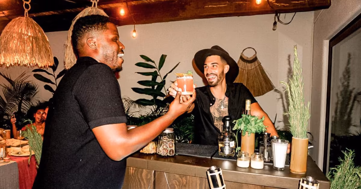 A smiling, tattooed bartender in a hat serves a cocktail to a happy customer at a lively, plant-filled bar with warm lighting.