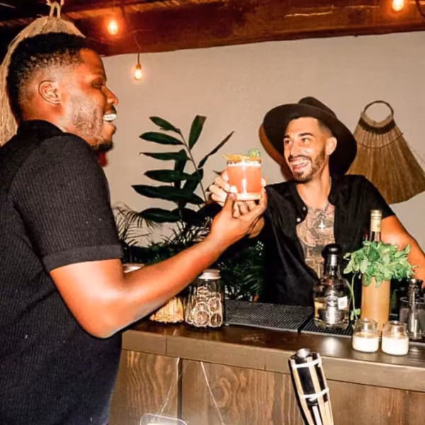 Two men smile and toast with cocktails at a cozy bar adorned with plants and warm lighting. One wears a hat behind the bar.