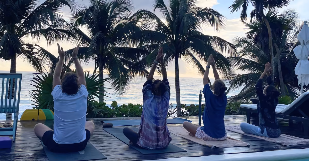 Four people do yoga on mats with arms raised on a deck by the ocean, surrounded by palm trees at sunrise or sunset.