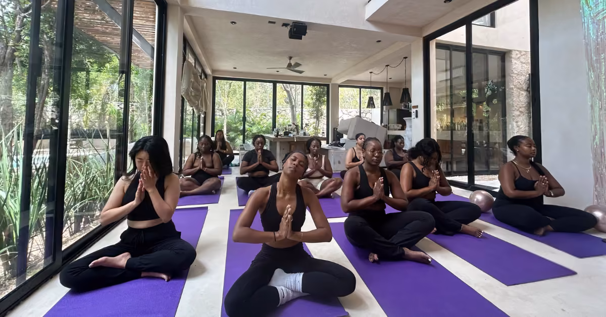 Women in black workout clothes sit cross-legged on purple mats, eyes closed, hands together, in a bright room with large windows.
