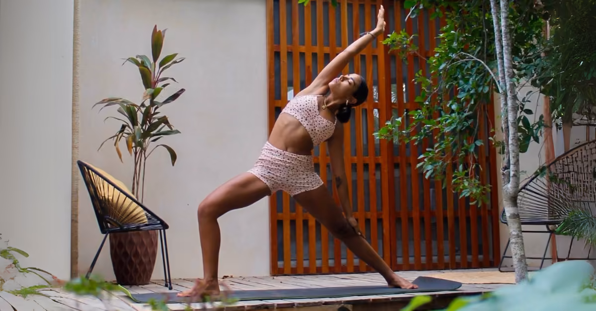 A woman practices a side angle stretch on a yoga mat outdoors, wearing a matching set, amid lush plants and a wooden screen.