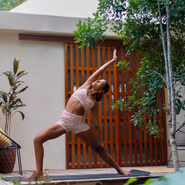 A woman in pink workout clothes practices yoga on an outdoor mat, surrounded by greenery and modern wooden structures.