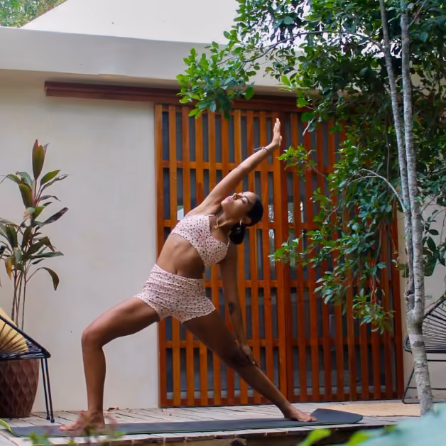 A woman in pink workout clothes practices yoga on an outdoor mat, surrounded by greenery and modern wooden structures.