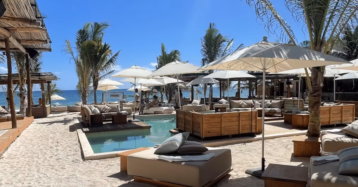 Outdoor beach lounge at Rosa Negra Beach Club with cushioned sofas, wooden tables, and umbrellas by a small pool on sandy ground. Palm trees and cabanas surround the area, with ocean views under a clear blue sky.