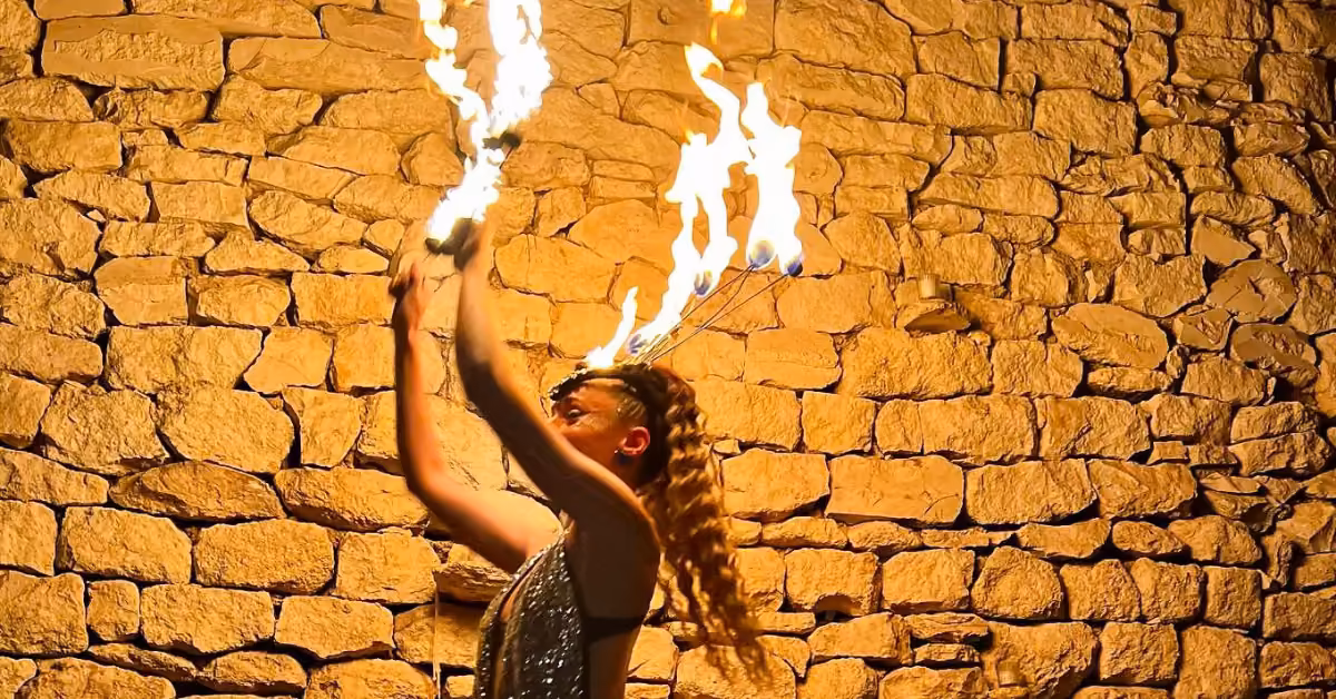 A performer with long braids lifts flaming torches above their head, entertaining guests in front of a rustic stone wall.
