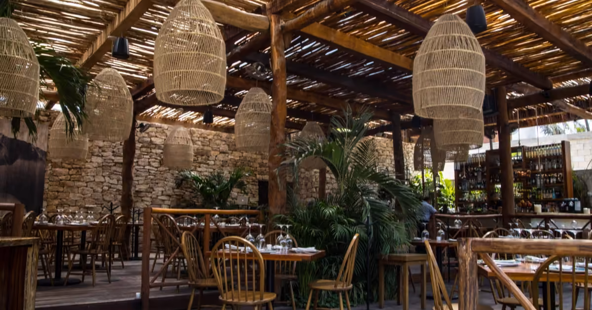 Outdoor restaurant with wooden tables and chairs, wicker pendant lights, green plants, stone walls, and a pergola roof.
