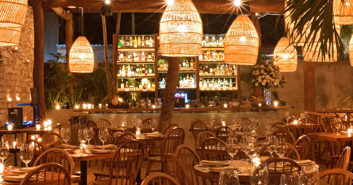 A cozy, warmly lit restaurant with wooden tables, wicker lights, glassware set neatly, and a bottle-lined bar in the background.