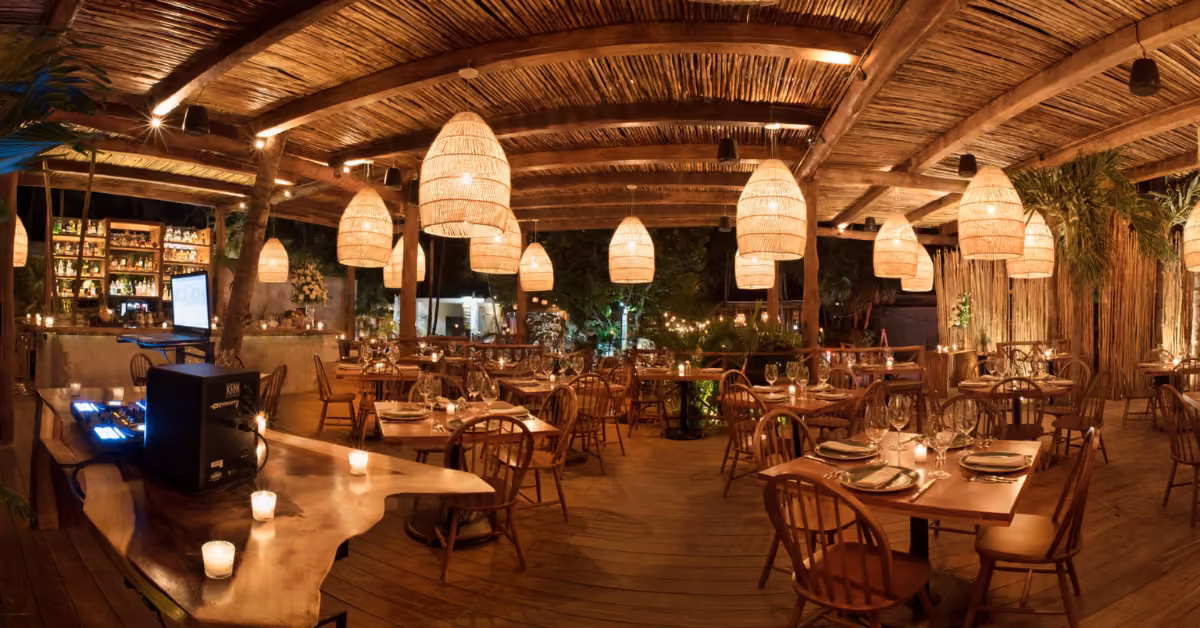 Warmly lit restaurant with wooden tables, woven lights, rustic decor, and greenery creating a relaxed, inviting atmosphere.