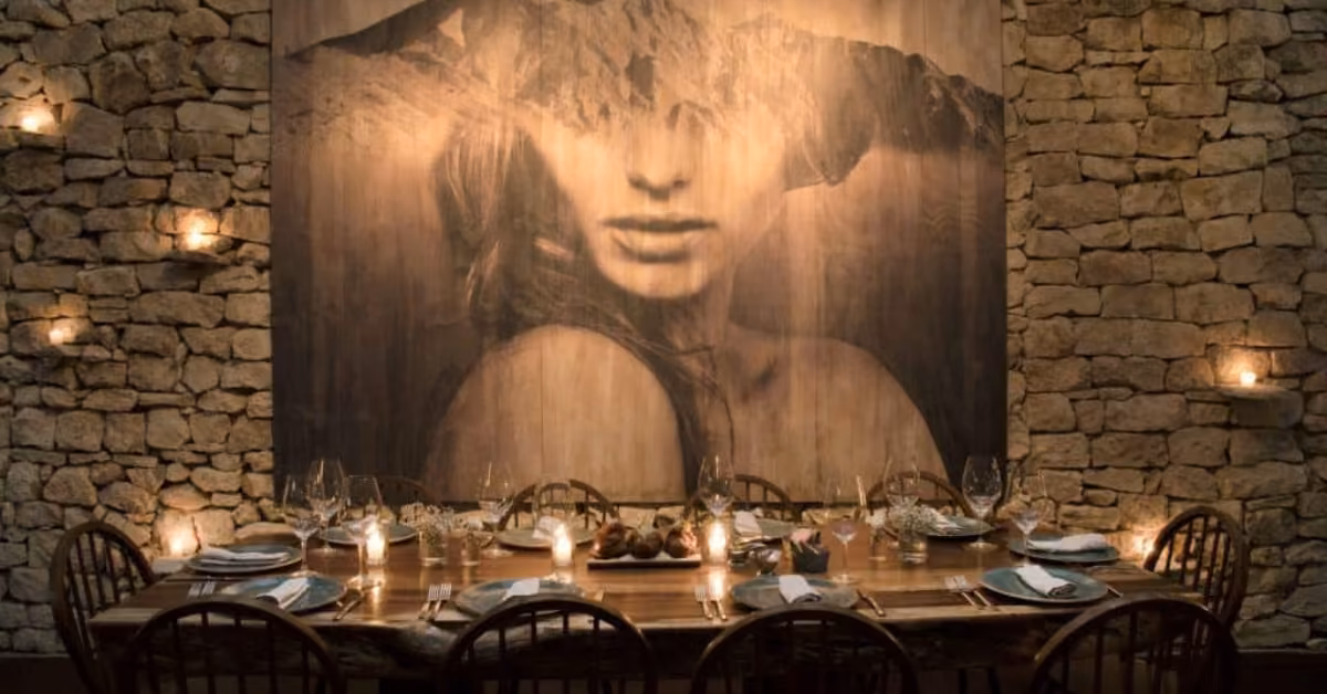 Cozy rustic dining room with a stone wall, wooden table, striking art of a woman’s face and mountains, and soft candlelight.