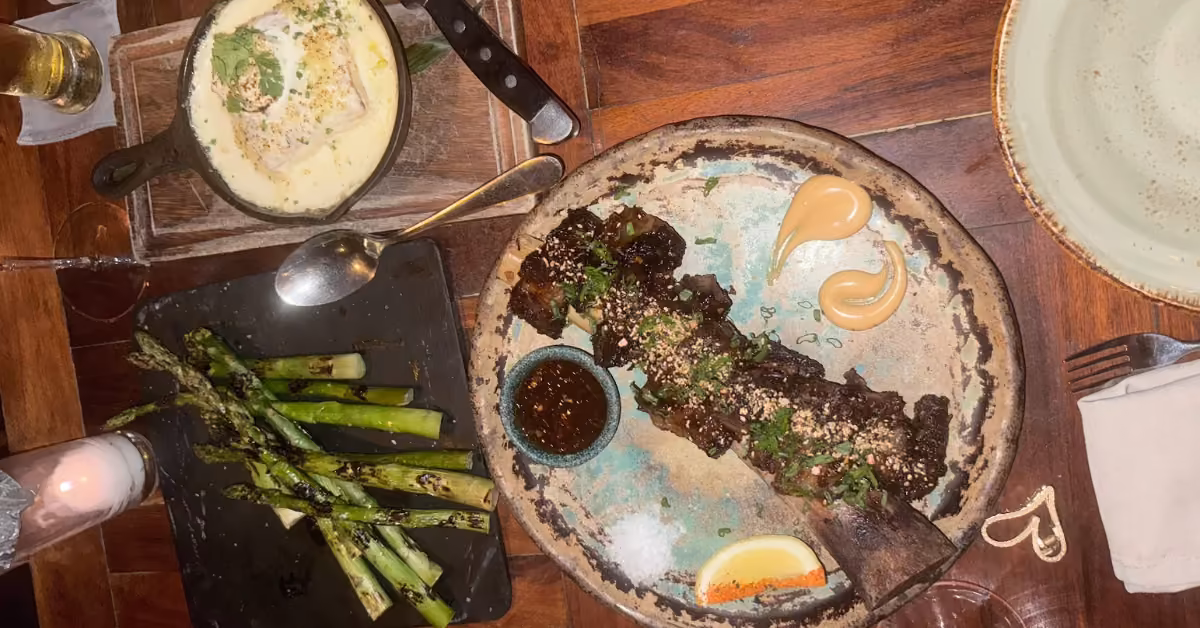 A rustic table at Rosa Negra shows grilled asparagus, short rib with herbs and sauce, mashed potatoes, and nearby utensils.