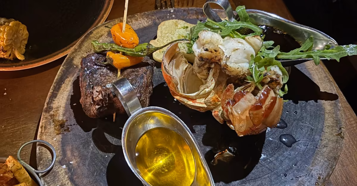 Grilled lobster tail, steak, cherry tomatoes, arugula, corn cob, and sauce beautifully arranged with char marks and garnish.