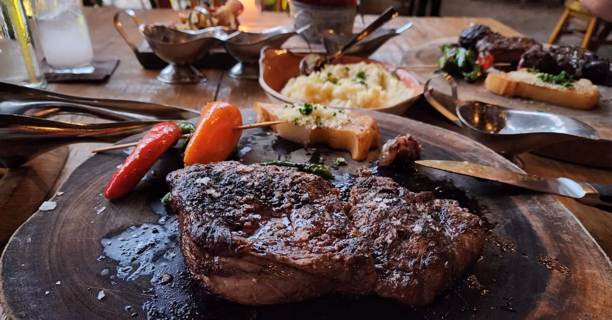 Grilled steak with salt on a wooden platter, served with roasted veggies, mashed potatoes, bread, and sauces on a rustic table.