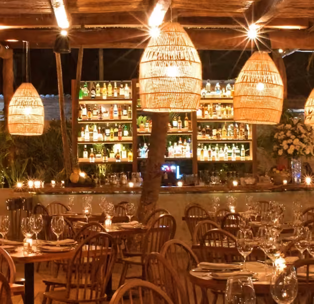 A cozy restaurant with wooden tables, woven lamps, and a stocked bar creates a warm, inviting atmosphere for dining.