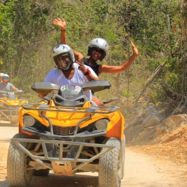 Two helmeted riders smile and wave on a yellow ATV along a dusty forest trail, with another ATV following among the trees.